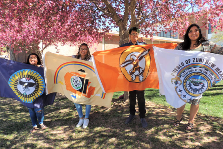 Four students standing under trees with native tribal flags